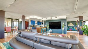 Un salon moderne avec un canapé d’angle gris, un coin cuisine doté de meubles bleus et un coin repas avec des chaises roses.