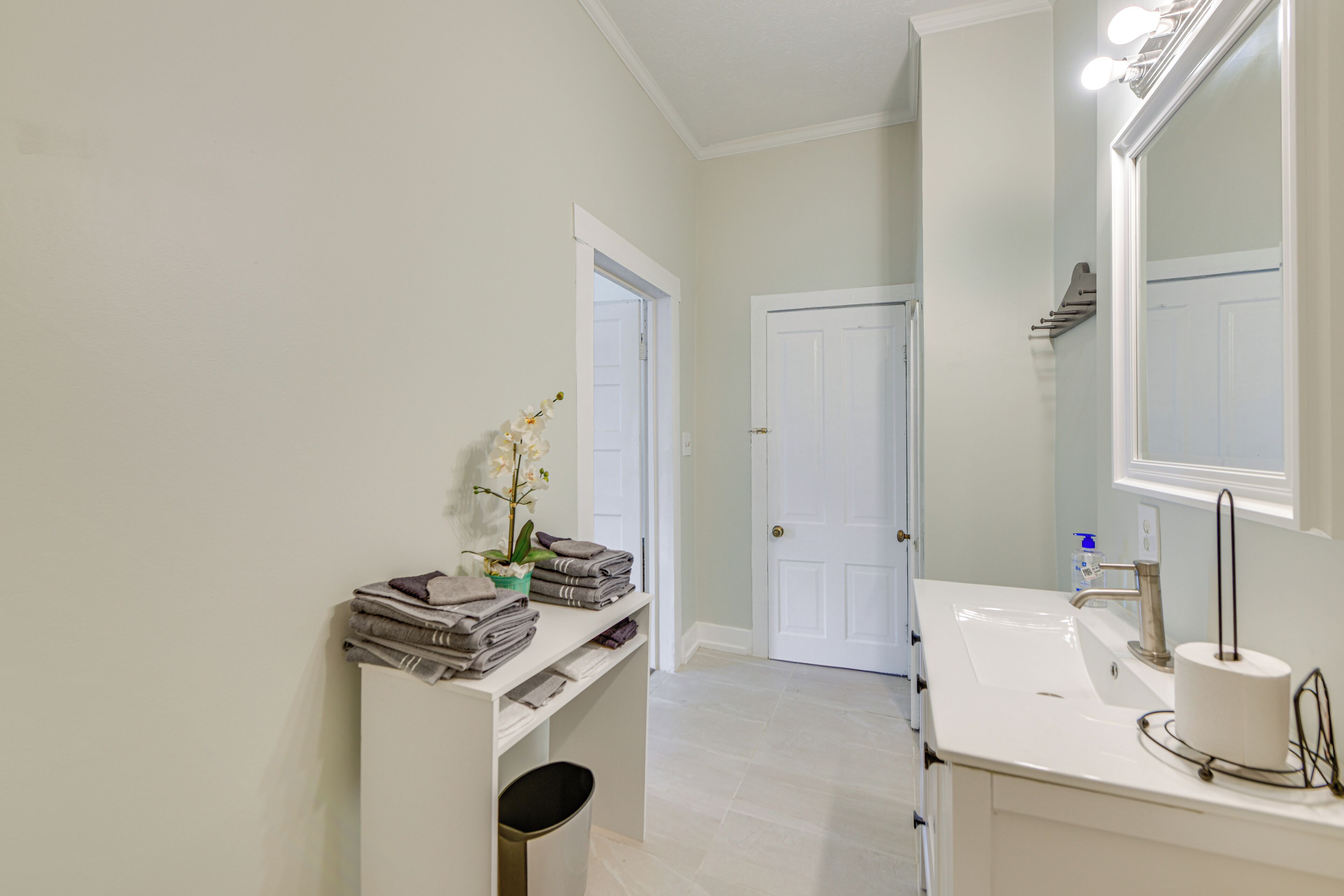 A bathroom with a white sink, a mirror, and a door. There is a shelf with folded towels and a potted plant.