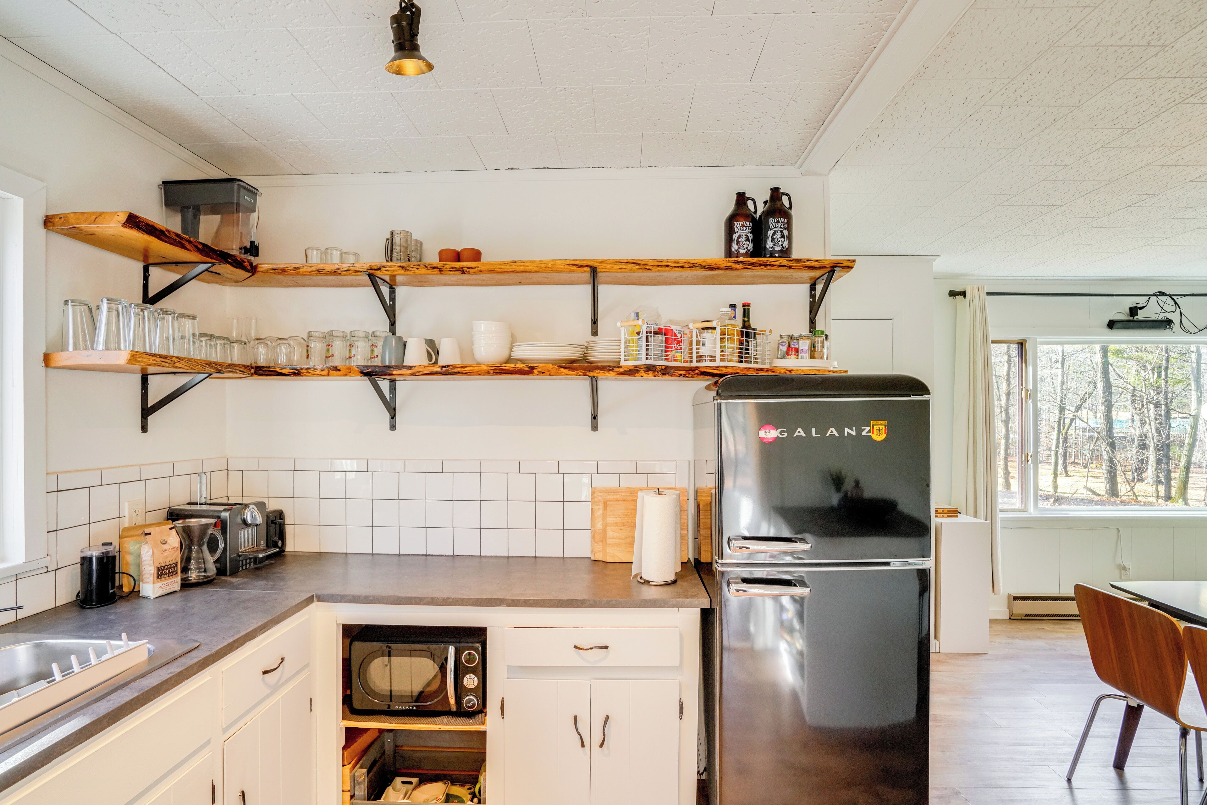 A modern kitchen with wooden shelves, a refrigerator, a microwave, and a dining area with chairs.