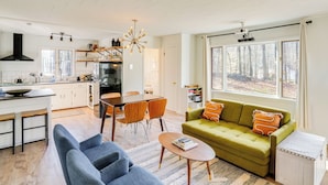 A modern kitchen with white cabinets, a dining area with wooden chairs, a green sofa with orange pillows, and large windows with a forest view. - Mtn Views + On-site Hiking Trails: Catskills Cabin (Round Top)