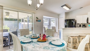 Uma sala de jantar com mesa posta para quatro pessoas, uma cozinha com geladeira e uma porta de vidro que se abre para um pátio externo.