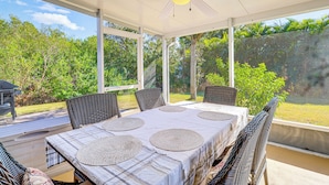 Um alpendre coberto com uma mesa posta para quatro pessoas, rodeado por cadeiras de vime, com vista para um jardim exuberante.