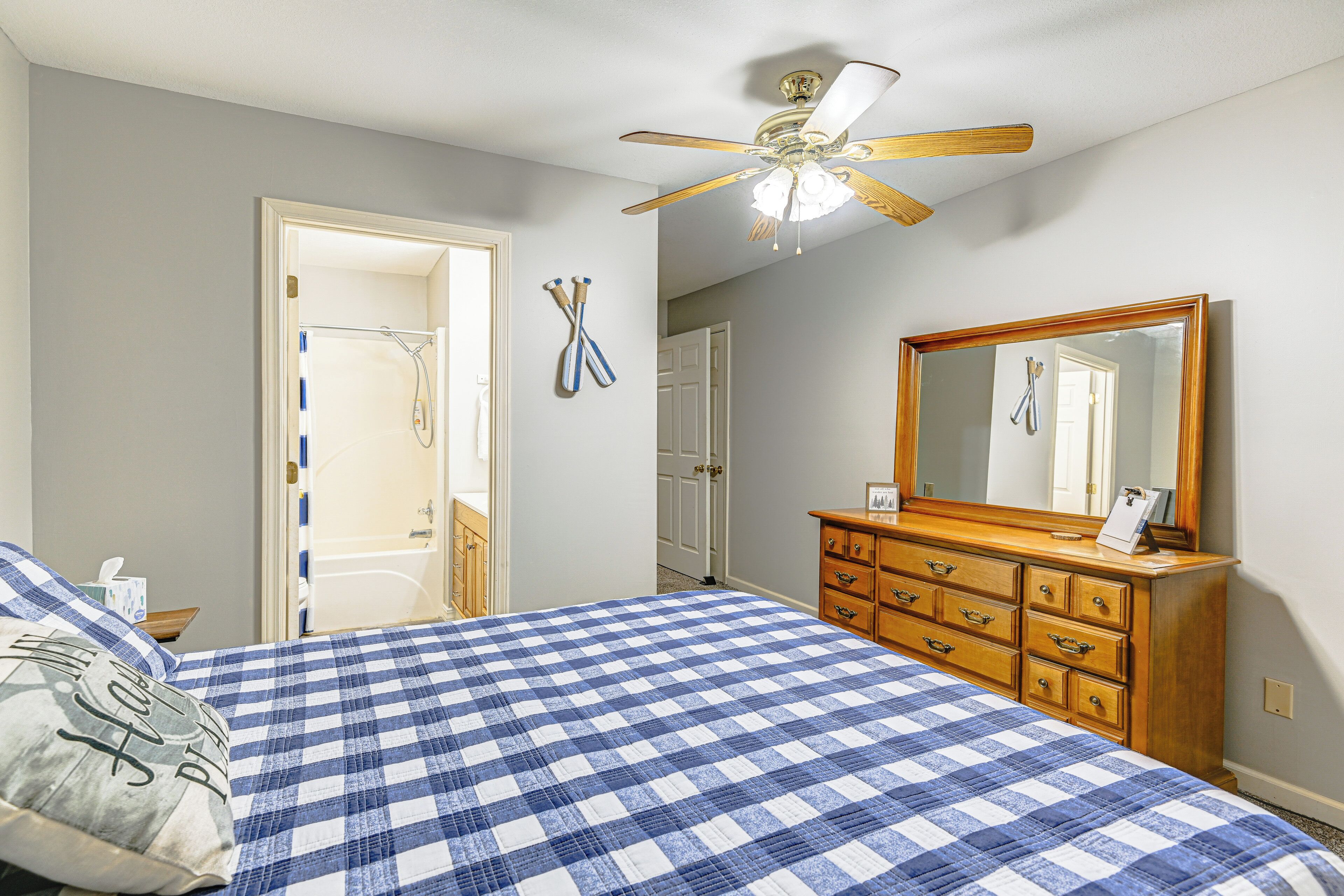 A bedroom with a checkered bedspread, a wooden dresser, a ceiling fan, and a mirror.
