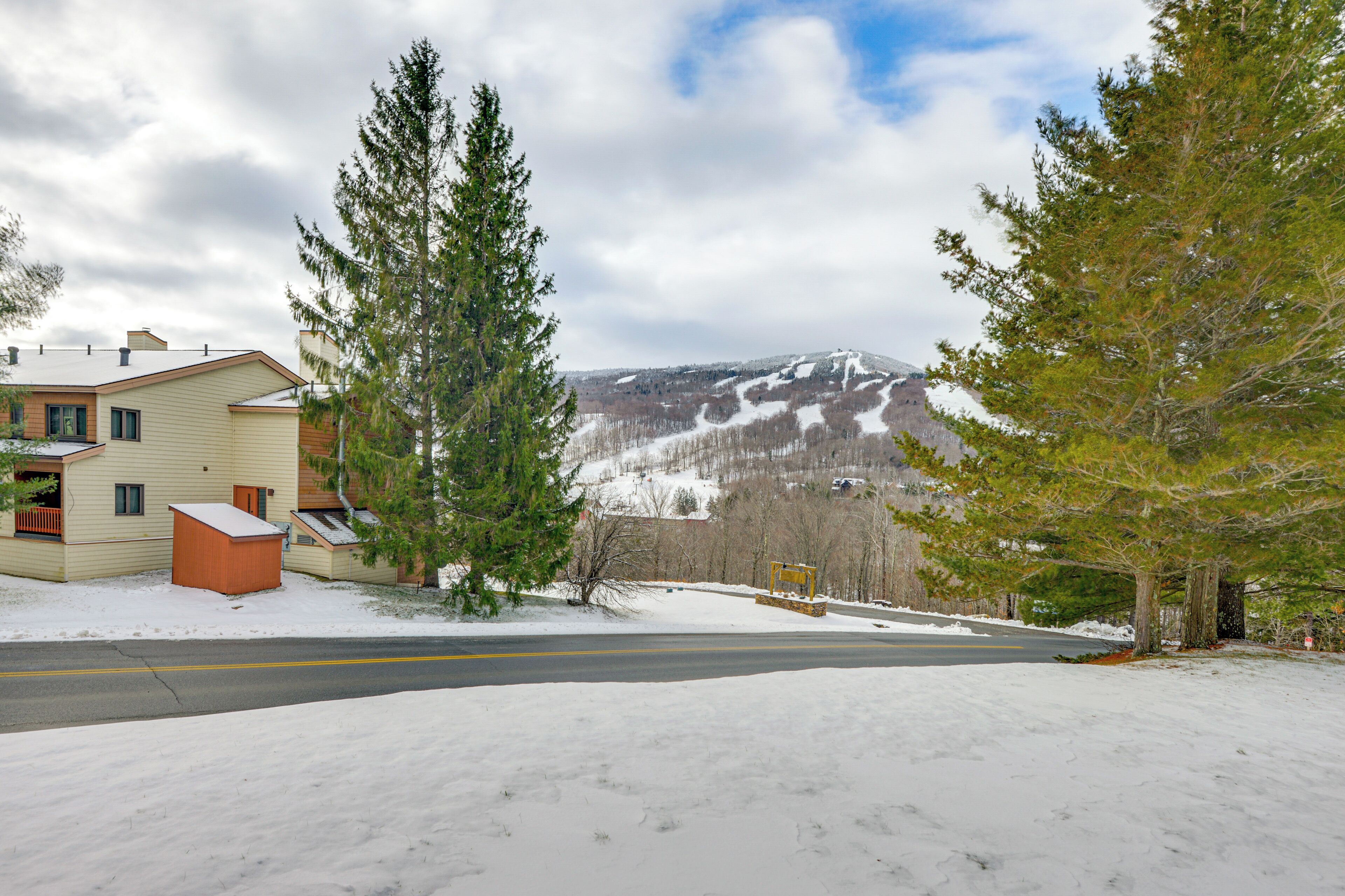 A residential area with a road, snow-covered ground, and a mountain in the background.