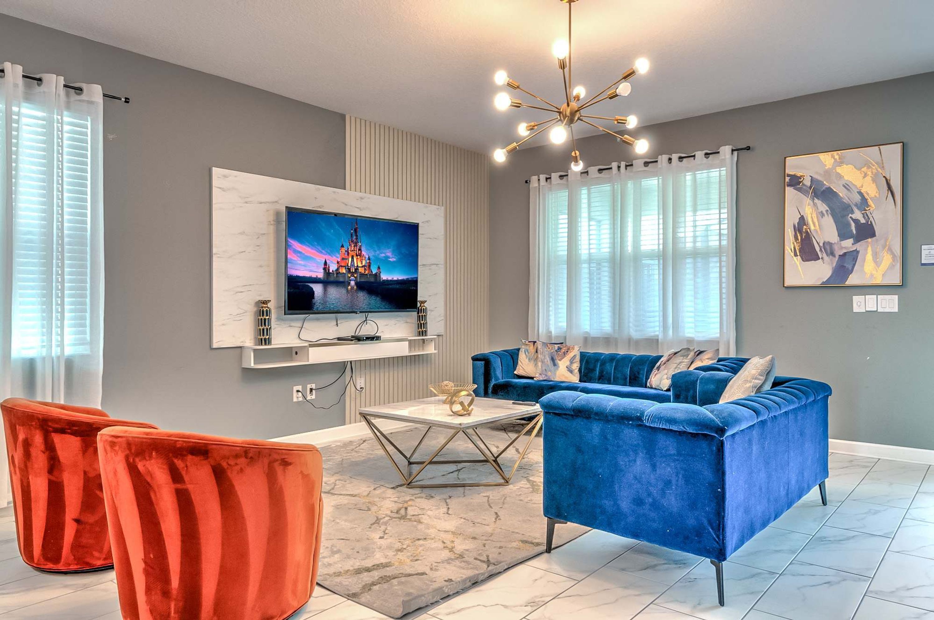 Ein modernes Wohnzimmer mit einem blauen Sofa, roten Sesseln, einem an der Wand montierten Fernseher und einer Pendelleuchte.