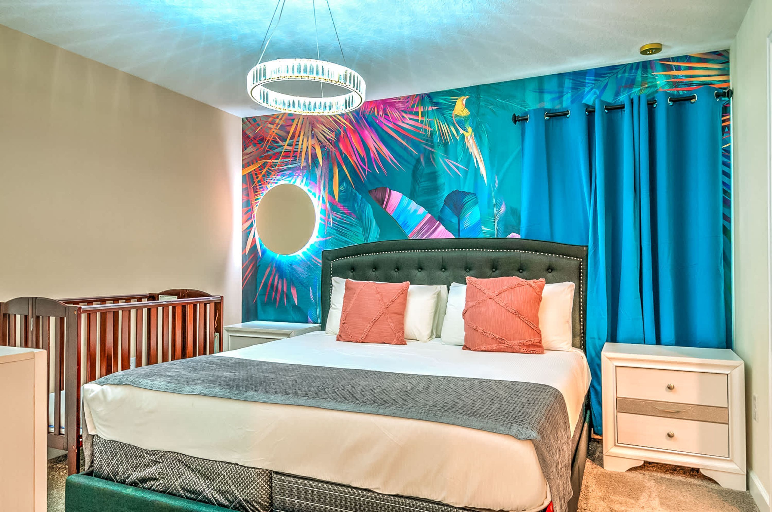 A bedroom with a large bed, a tufted headboard, a nightstand, a crib, and a vibrant tropical mural on the wall.