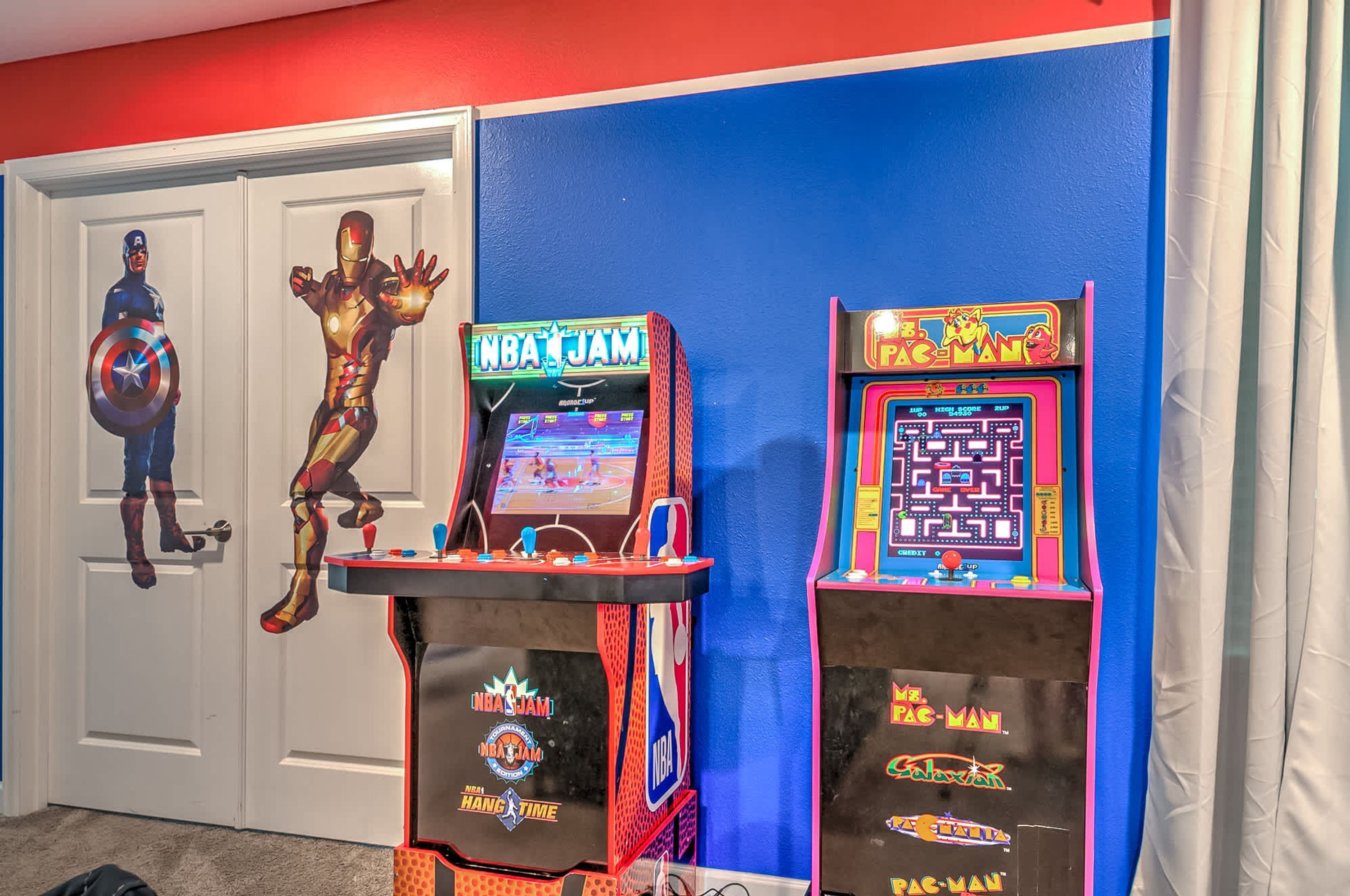 Two arcade machines labeled "NBA JAM" and "Pac-Man" in a room with red and blue walls.