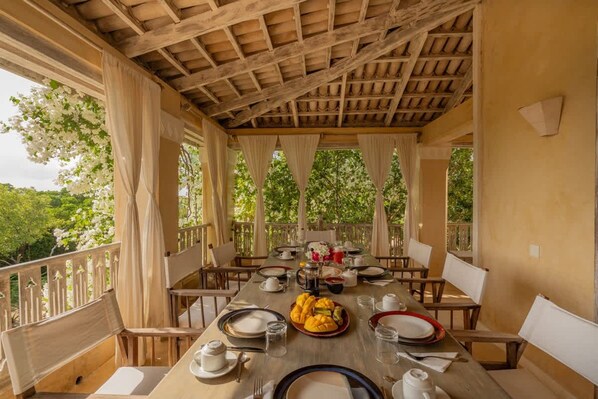 An outdoor dining area with a wooden table set for a meal, surrounded by chairs, and a view of greenery. - Amani House 5 Bedrooms Sleeps 10 (Manda Island)