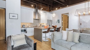 Un salon moderne avec un canapé gris, un canapé gris clair et une table basse en bois. On y trouve également un coin cuisine avec des meubles blancs, un réfrigérateur et un coin repas avec un tabouret de bar.