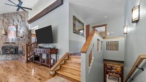 A spacious living room with a stone fireplace, wooden flooring, and a flat-screen TV mounted on the wall.