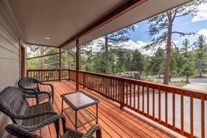 Una terraza de madera con mobiliario de mimbre y una mesa de vidrio, con vistas a un área boscosa y cielo despejado.