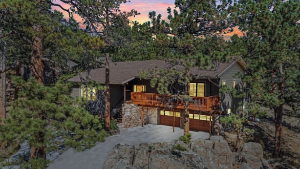 A house surrounded by dense pine trees, with a stone foundation and a wooden deck.