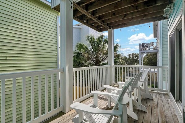 A covered porch with white railings and wooden deck, featuring outdoor seating and a view of a residential area with palm trees.
