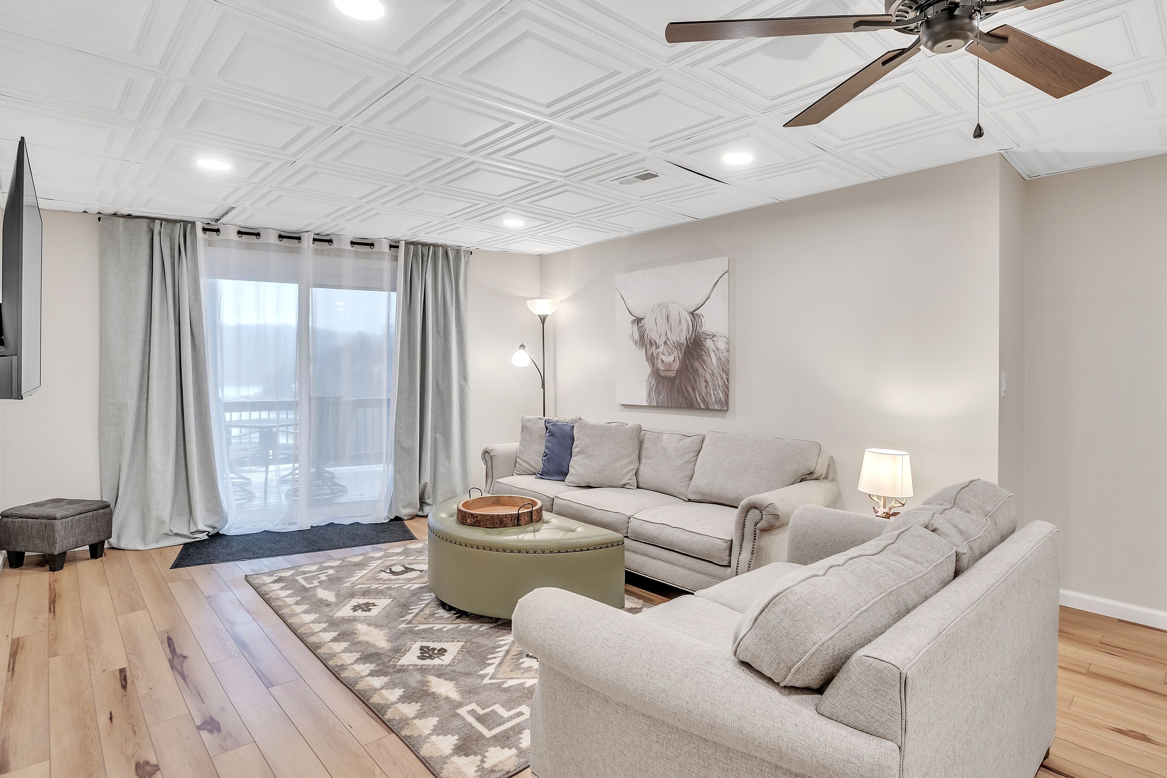 A modern living room with a ceiling fan, a sofa, an ottoman, and a large window with curtains.