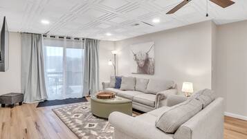 A modern living room with a ceiling fan, a sofa, an ottoman, and a large window with curtains.