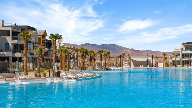 Piscina de un complejo turístico rodeada de palmeras y edificios modernos, con montañas al fondo.