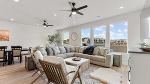 Una sala moderna con un sofá seccional, una zona de comedor con mesa de madera y grandes ventanales que ofrecen vistas del vecindario.