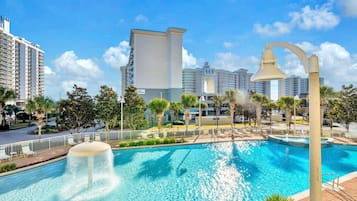 Una piscina con una fuente de agua, rodeada de palmeras y una valla, con edificios altos al fondo.