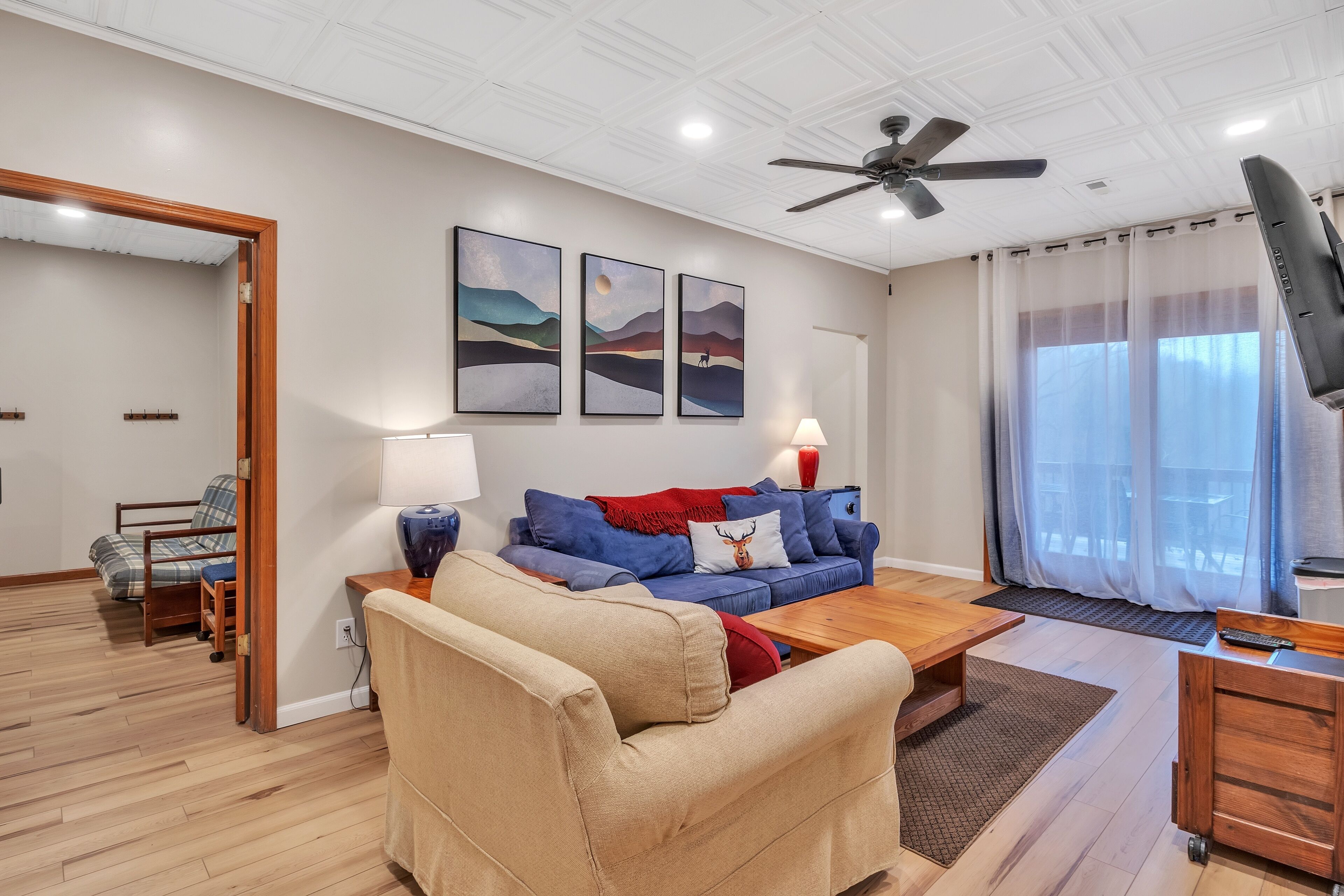 A living room with a beige sofa, a blue sofa, a wooden coffee table, a TV, and a ceiling fan. There are three framed pictures on the wall and a window with curtains.