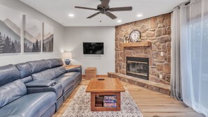 A modern living room with a stone fireplace, leather sofa, and a ceiling fan. - Deerfield Hideaway At Old Smokey 5 Bedroom Home (La Follette)