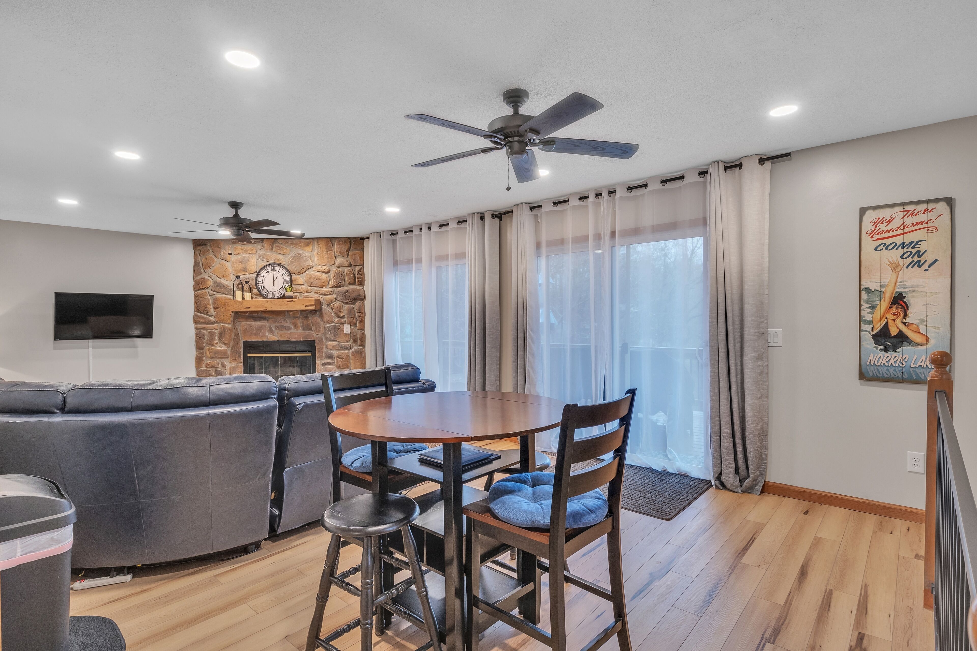 A modern living room with a stone fireplace, a round dining table with chairs, a sofa, and a ceiling fan.