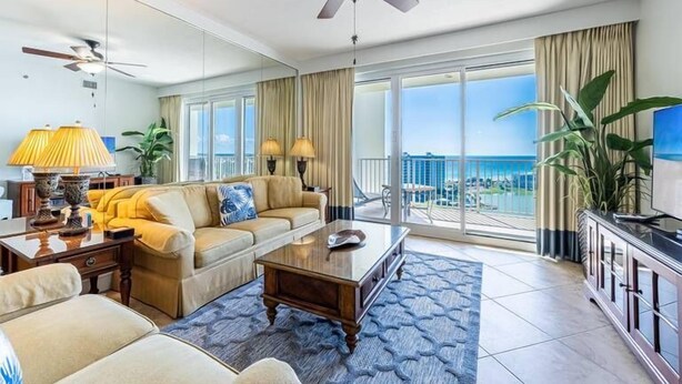 A spacious living room with a sofa, coffee table, and a TV on a cabinet. There are two ceiling fans, a large window with a view, and a balcony with curtains. - Spectacular view of the Gulf and just steps away from the pool and beach! (Miramar Beach)