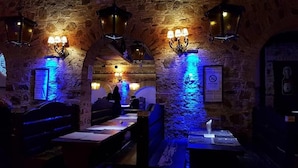Una habitación tenuemente iluminada con paredes de piedra, bancos y mesas de madera. Hay lámparas fijadas a la pared y un cartel con un símbolo de prohibición.