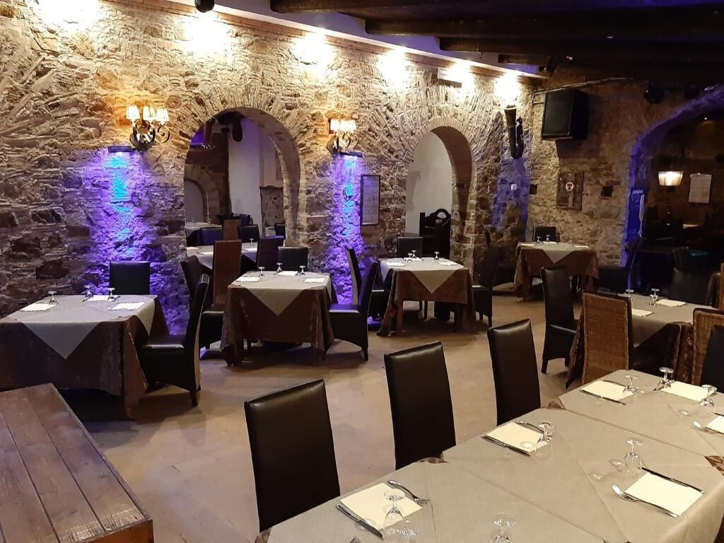 Ein Restaurant mit Steinwänden, Tischen gedeckt mit weißen Tischdecken und Stühlen, beleuchtet mit violettem Licht.