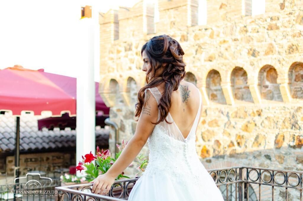 Una persona con un vestido blanco de pie junto a un muro de piedra con arcos, sosteniendo un ramo de flores.