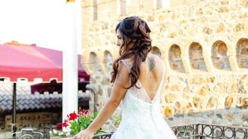 Una persona con un vestido blanco de pie junto a un muro de piedra con arcos, sosteniendo un ramo de flores.
