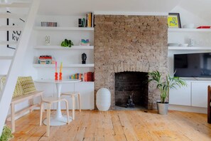 Ein Zimmer mit Kamin aus Ziegelsteinen, Holzboden, einem weißen runden Tisch mit Stühlen und einem Bücherregal mit Büchern und Dekoration.