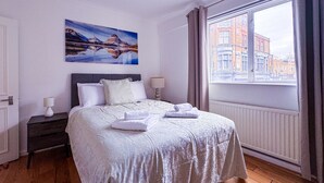 Habitación de hotel con cama, mesita de noche, lámpara, un cuadro y ventana con vistas a edificios.