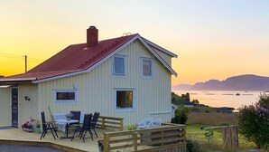 A single-story house with a porch, a dining area with chairs and a table, and a view of the water and hills in the background. - 4 Person Holiday Home in Alsvag (Oksnes)