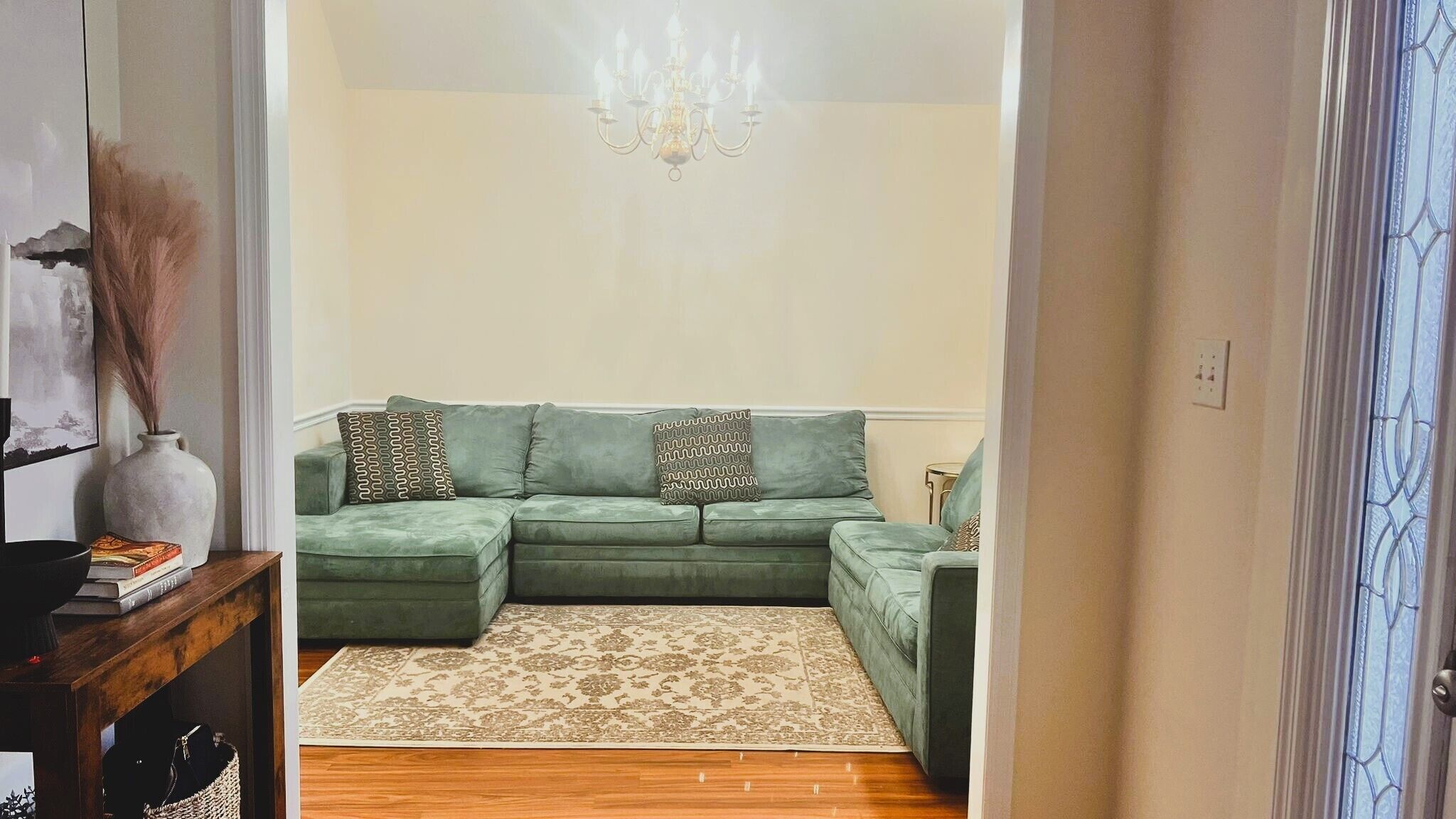 A green sectional sofa with patterned pillows, a beige patterned rug, and a chandelier.