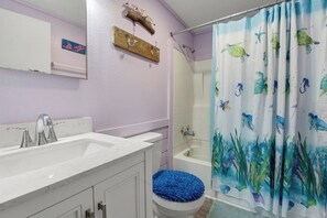 Une salle de bain avec un lavabo blanc, un miroir, une étagère en bois avec des crochets, un tapis de bain bleu, un rideau de douche orné de motifs marins et des toilettes blanches.