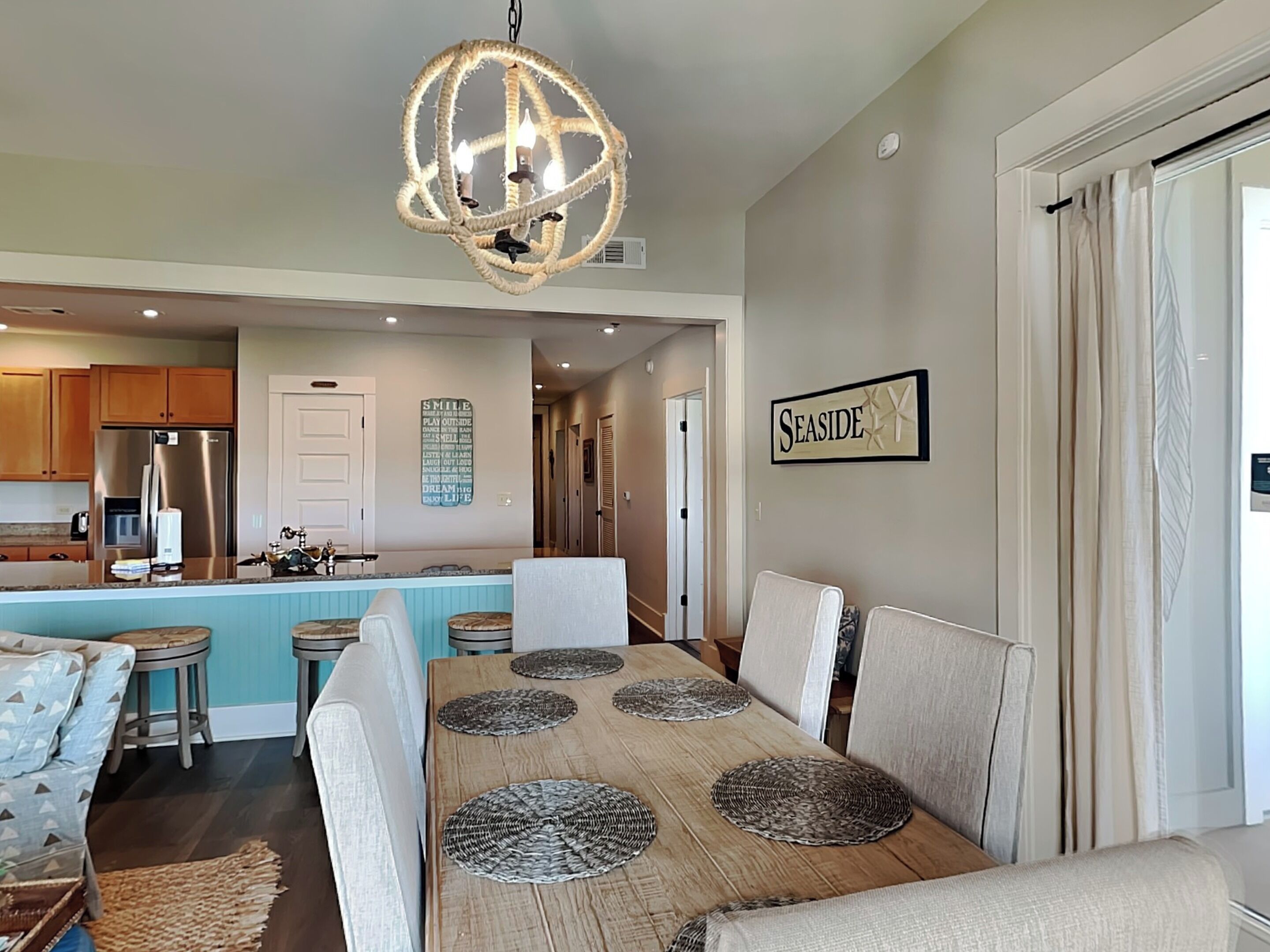 A modern kitchen with stainless steel appliances, a central island with bar stools, and a dining area with a wooden table and woven placemats.