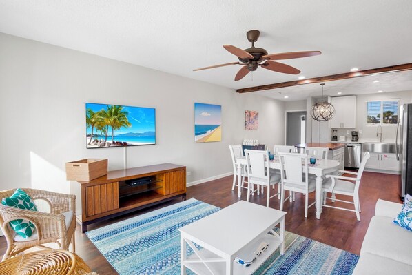 A spacious living room with a large TV, a ceiling fan, and a dining area with white furniture.