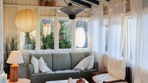 A cozy room with a grey sofa, a wooden chair, a table with a vase, and a lamp. There are windows with curtains and a ceiling fan. - 6 Person Holiday Home in Sondeled (Risør)