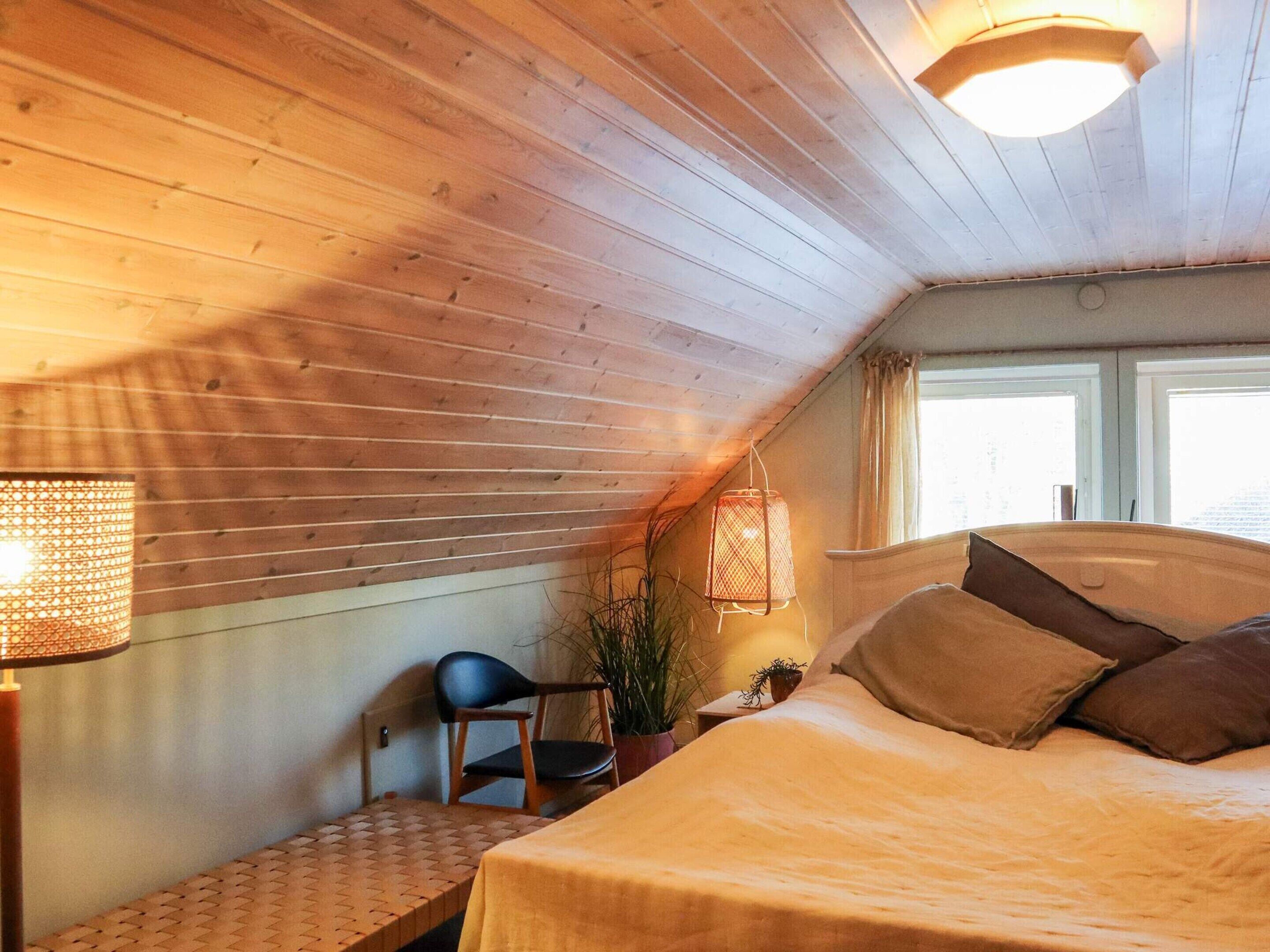 Una habitación acogedora con techo inclinado, paredes de madera, una cama con almohadas, una silla, una mesa y una ventana con cortinas.