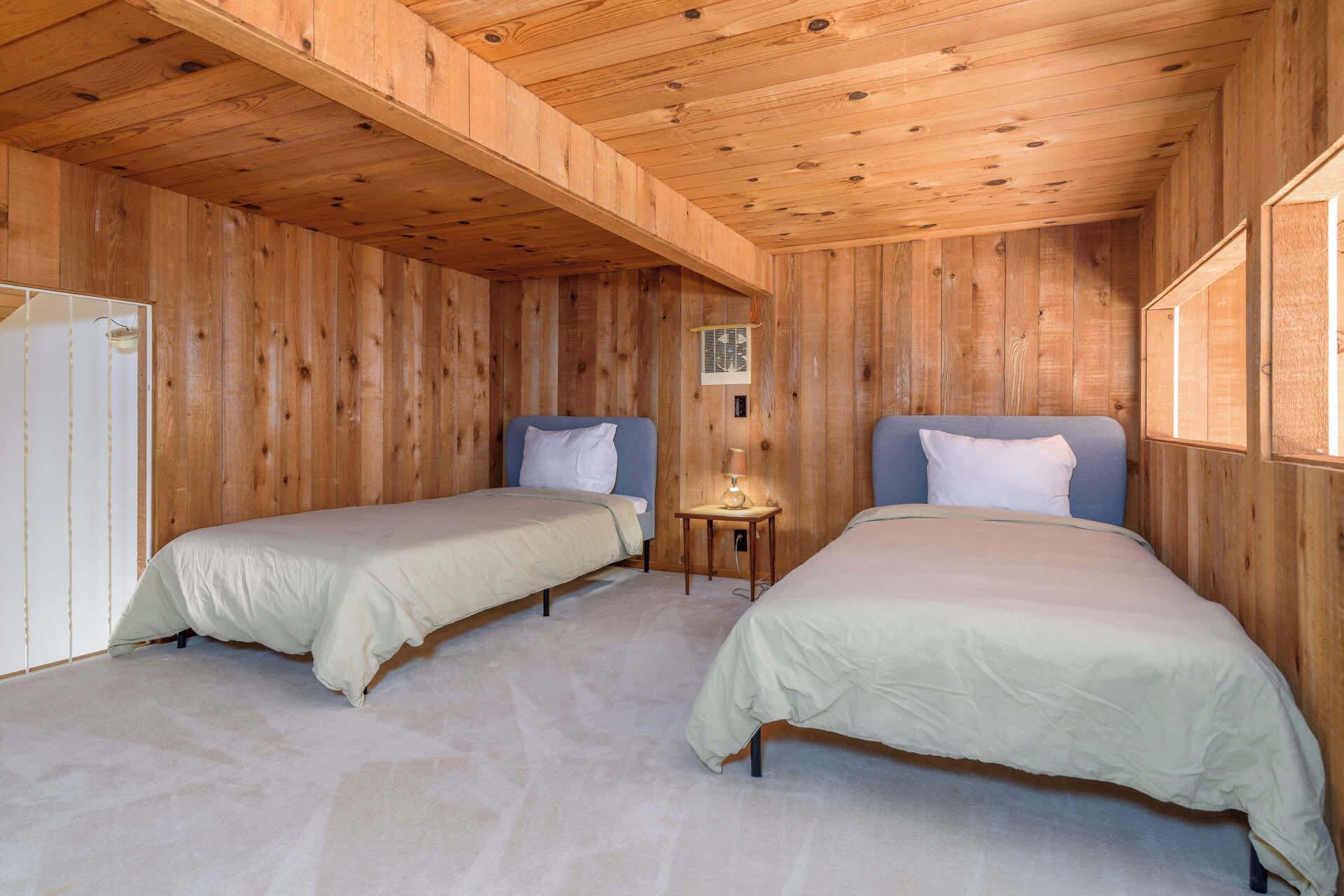 A room with two single beds, wooden walls, a small table with a lamp, and a window.