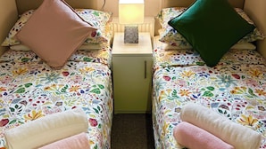 Une petite chambre avec deux lits, chacun agrémenté d’une literie à motifs floraux, une table de chevet avec une lampe et une vue sur un champ verdoyant par la fenêtre.