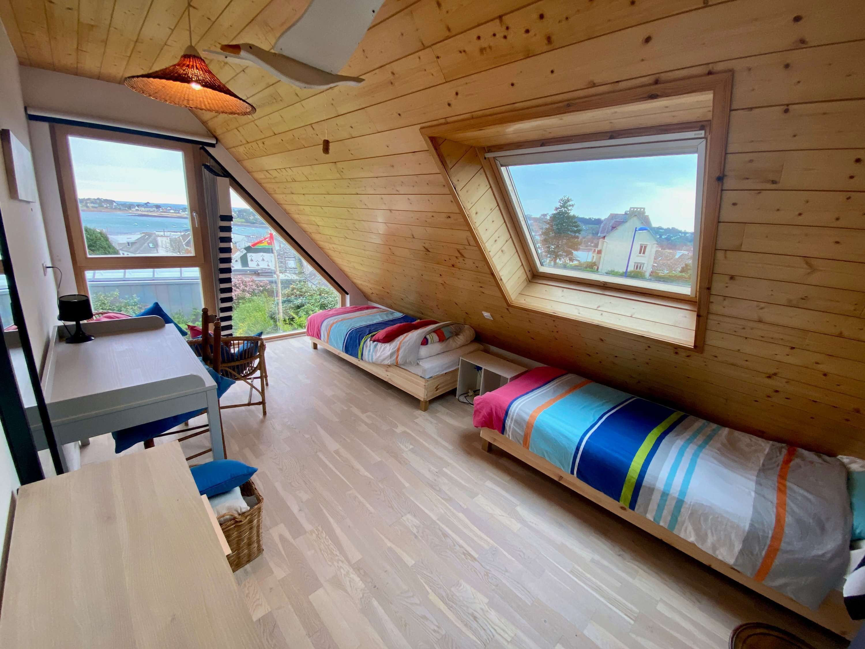 Une chambre confortable située sous les toits, avec un plafond en pente, une grande fenêtre donnant sur l’extérieur, un bureau en bois et un lit agrémenté d’une literie aux couleurs vives.