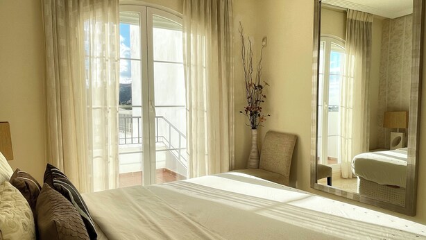 A hotel room with a large bed, a chair, a vase with branches, and a balcony with a railing.