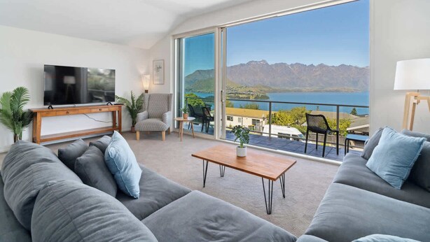 A modern living room with a large TV, grey sofas, a wooden coffee table, and a balcony with a view of mountains and a lake.