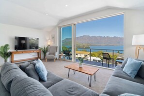 Una sala de estar moderna con un televisor grande, sofás grises, una mesa de centro de madera y un balcón con vistas a montañas y un lago.