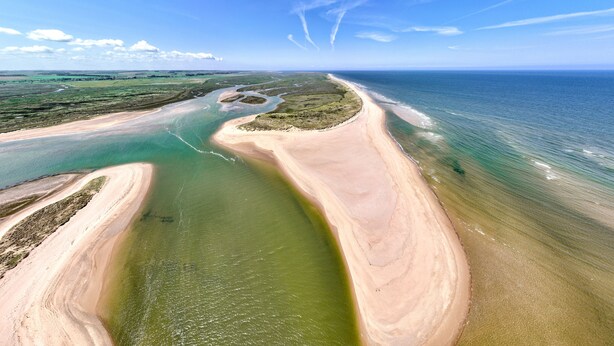 Une vue aérienne d’un paysage côtier, avec une rivière sinueuse, des plages de sable et un ciel bleu dégagé.