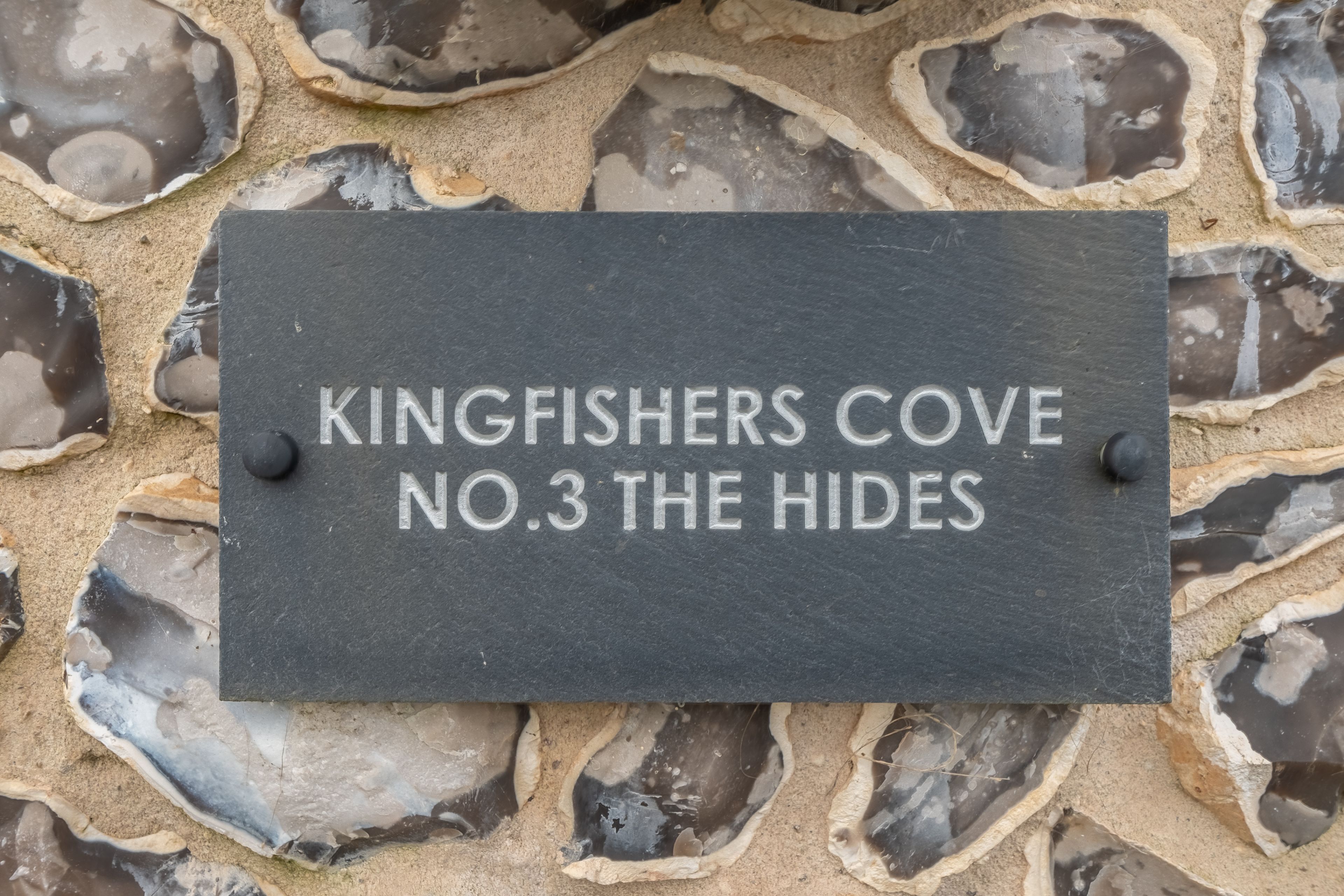A sign reading "Kingfishers Cove No.3 The Hides" mounted on a stone wall.