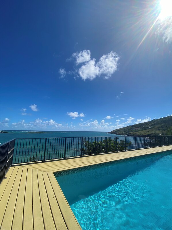 Una terraza de madera que da a una piscina azul cristalina, con vista al océano y a una costa distante.