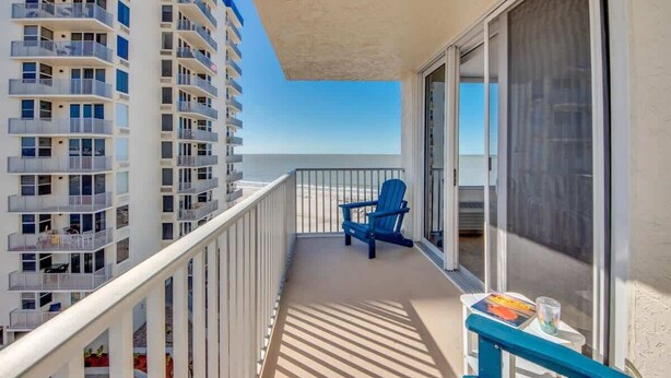 Ein Balkon mit blauem Stuhl, einem kleinen Tisch mit Buch und Glas sowie Blick auf das Meer und angrenzende Gebäude.