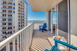 Un balcon avec une chaise bleue, une petite table sur laquelle se trouvent un livre et un verre, et une vue sur l’océan et les bâtiments adjacents.